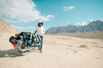 Mid adult woman with motorcycle standing on desert against blue sky during sunny day