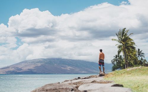 Rear view of shirtless man standing on rock against sky