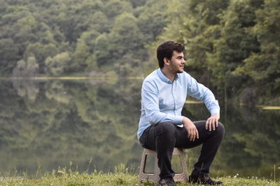 Young man looking away while sitting on stool against lake in forest