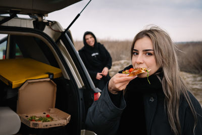 Portrait of young woman eating pizza and standing by car on the beach