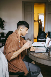 Side view of male freelancer having coffee while doing video call with colleague on laptop at home office