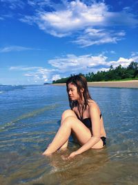 Portrait of young woman in bikini sitting on sea against sky