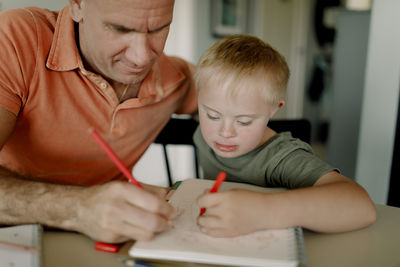 Father and son with down syndrome drawing together in book at dining table