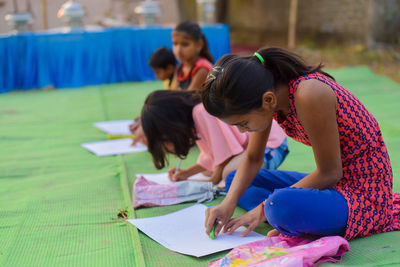 Side view of girls drawing on paper while sitting outdoors