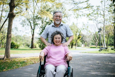 Portrait of smiling senior woman by man sitting on wheelchair at road in park