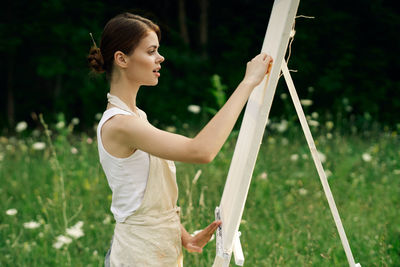 Side view of young woman holding plant while standing on field