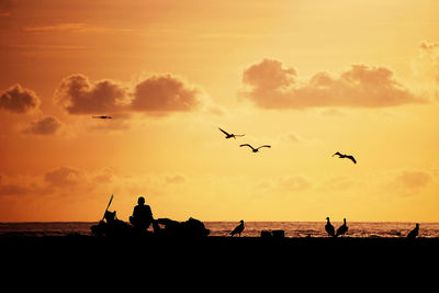Silhouette birds flying over sea against orange sky