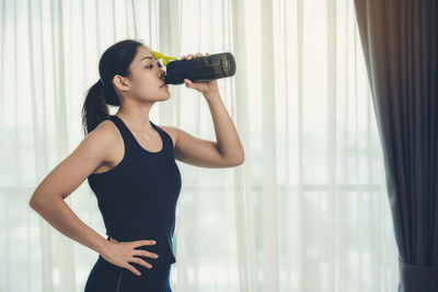 Young woman drinking glass while standing by window