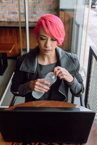 Stylish young female with dyed hair in black outfit sitting at table and having bottle of water while looking at laptop screen in cafe