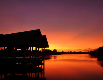 Silhouette houses by lake against romantic sky at sunset