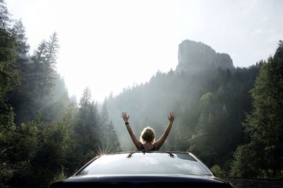 Rear view of woman with arms raised in car at forest against sky