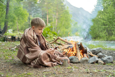 Boy wrapped in blanket using smart phone in forest