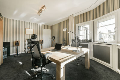 Wooden table with laptop and comfortable armchairs placed near panoramic window in spacious room with striped wallpaper in contemporary apartment