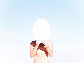 Shirtless man covering face with ellipse against sky