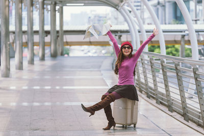 Portrait of smiling mid adult woman sitting on suitcase at elevated road