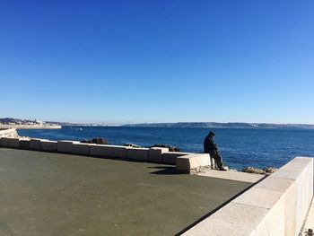 Man standing on retaining wall by sea against clear blue sky