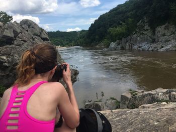 Woman photographing on rock by river against sky