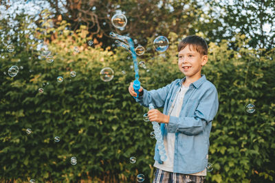 A boy is playing with soap bubbles in a summer park, among the greenery.