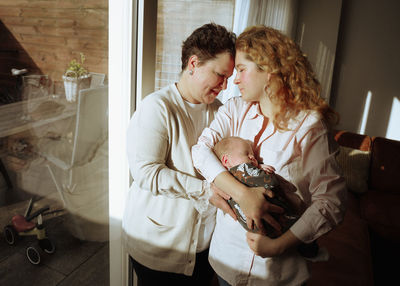 Side view of mother and daughter standing in front of window at home and holding a newborn son