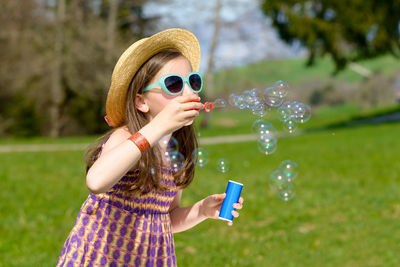 Girl wearing sunglasses and hat blowing bubbles while standing at park