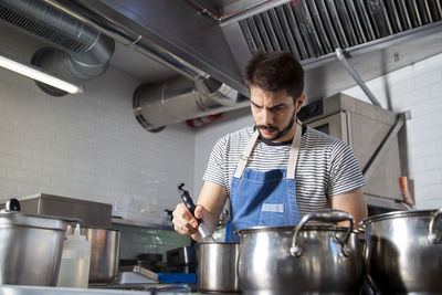 Concentrated young bearded hispanic male chef in casual clothes and apron stirring boiling dish with whisker in saucepan while cooking in restaurant kitchen in daytime