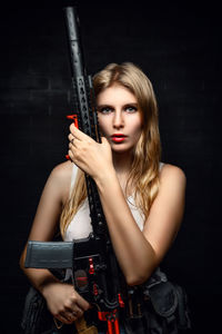 Portrait of young woman holding camera while standing against black background