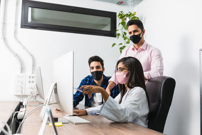 Anonymous entrepreneur in mask showing project on desktop computer while interacting with young coworkers in office