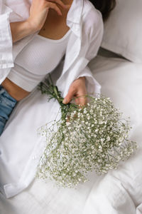 Woman wearing a white shirt and blue jeans is holding a bouquet of white flowers on a bed