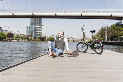 Smiling mature man with laptop and bicycle sitting at the riverside in the city
