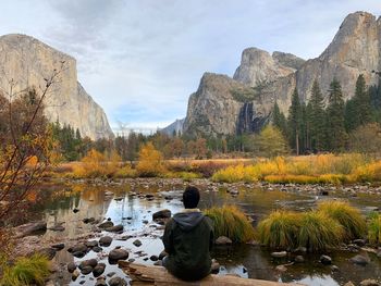 Rear view of man siting on driftwood against rock mountains