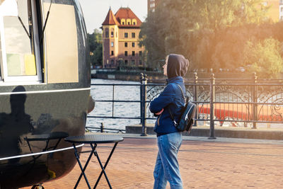 Young woman in hood and with backpack stands near food truck and reads menu. 