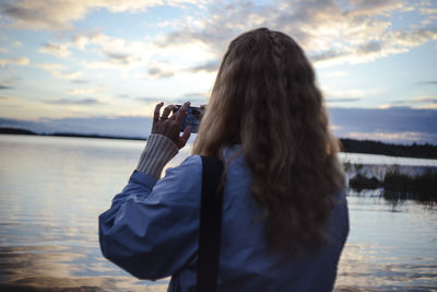 Rear view of woman photographing lake through smart phone at sunset