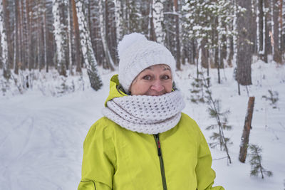 Senior woman in yellow jacket, white knitted hat and scarf looking at camera in winter snowy forest