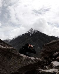 Hiker drinking water sitting on rocky mountain against sky