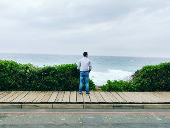 Rear view of man fishing on retaining wall by sea against sky