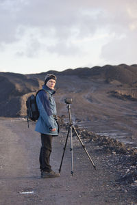 Portrait of man standing by digital video camera with tripod by field against sky