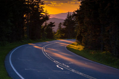 Empty road amidst trees in forest against sky at sunset