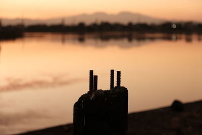 Close-up of silhouette wooden post by lake against sky during sunrise