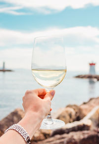 Woman hand with white wine glass on a sea, stones and lighthouse background. nice, french riviera