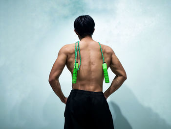 Rear view of shirtless young man with jumping rope standing against wall