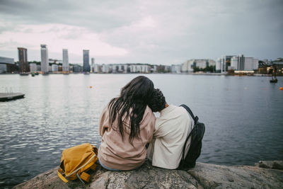 Rear view of teenage boy leaning on female friend while sitting on rock near sea in city