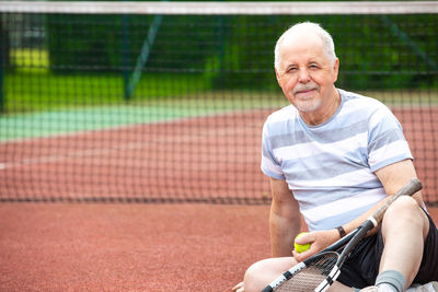 Portrait of smiling man holding tennis ball sitting on court