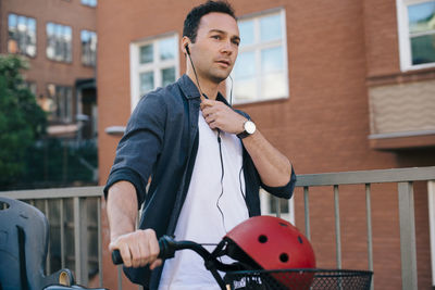 Mid adult man adjusting in-ear headphones while walking with bicycle against building in city