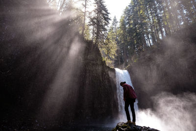 Low angle view of man standing on rock against waterfall in forest