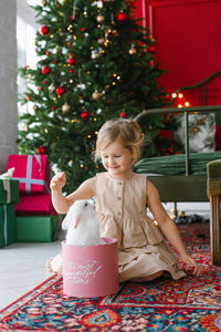 A little girl in a dress sits on the floor by the bed and looks at a rabbit sitting in a box