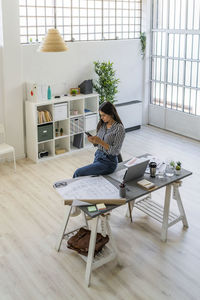 Young businesswoman using smart phone while sitting by blueprint at desk in creative workplace