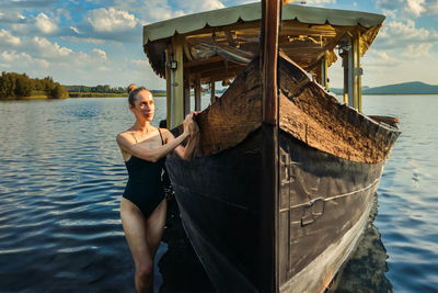 Slim attractive woman in one-piece swimsuit relaxing by lake and standing leaning near wooden ship
