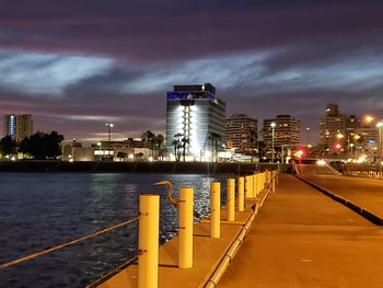 Illuminated bridge over river by buildings against sky at dusk