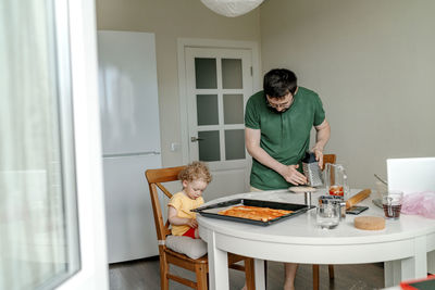 Man making pizza by daughter sitting on chair in kitchen