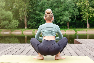 A slender woman sitting on a wooden platform by a pond on green mat in summer, does yoga